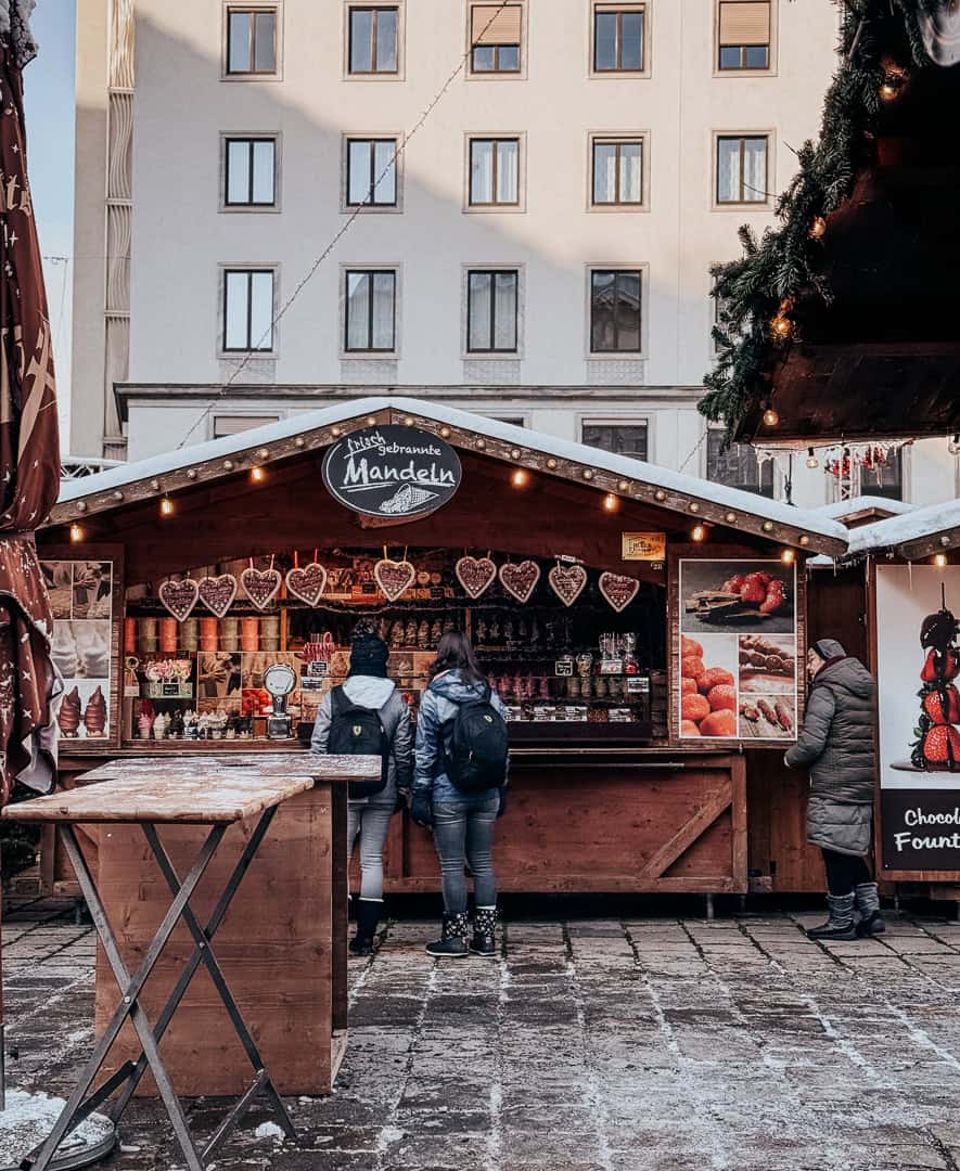 Visitors browse handcrafted heart-shaped decorations at a Vienna Christmas market stall, with architecture in soft focus in the background