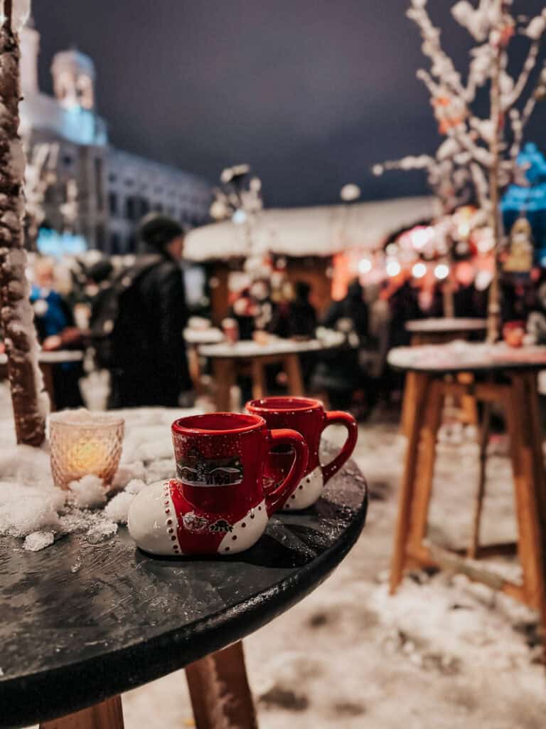 Red and white Christmas mugs resting on a snowy table at a Vienna market, with the cheerful hustle of the market and historical buildings in soft focus behind.