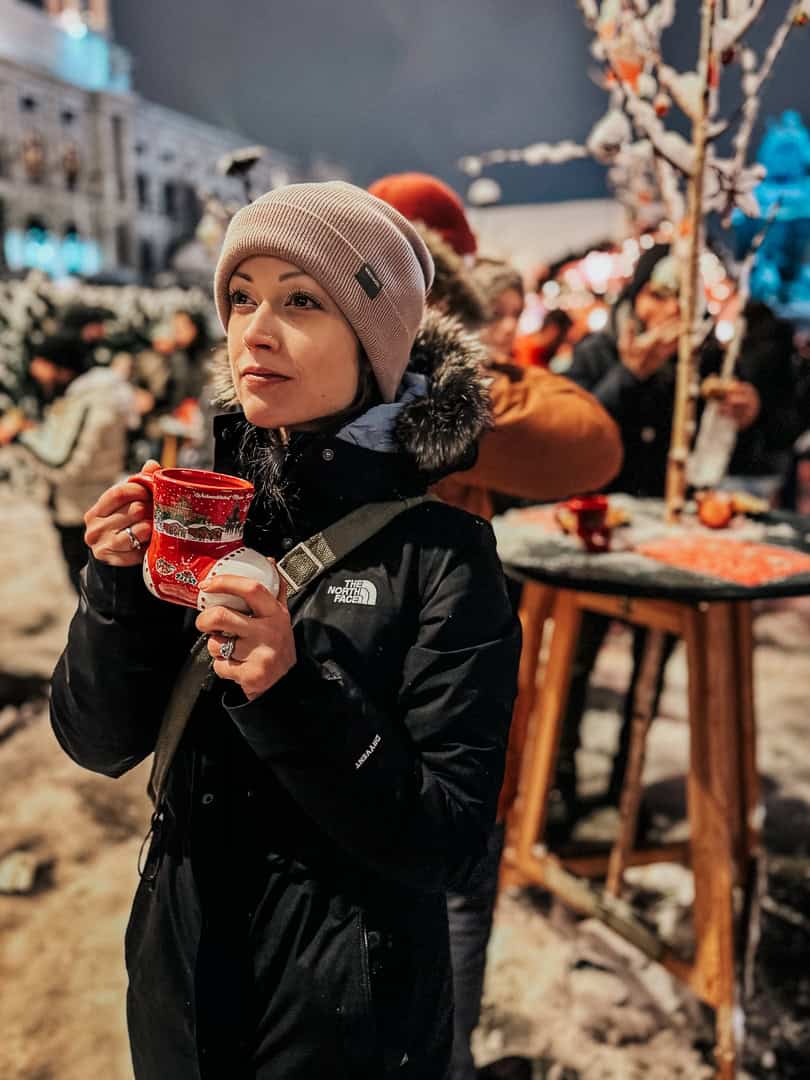 A person in winter attire enjoys a hot drink from a festive mug, standing amidst the lively atmosphere of a Vienna Christmas market at night.