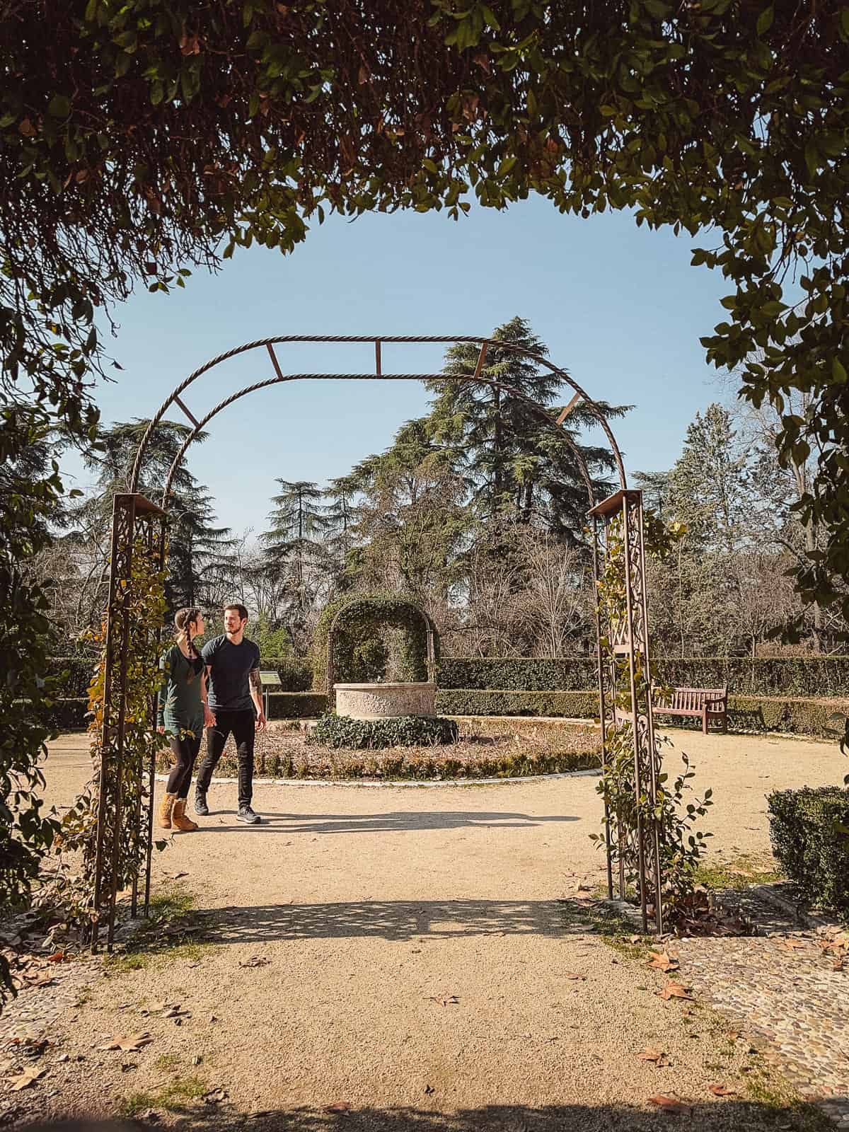 Two individuals walking under a leafy metal archway on a gravel pathway in the peaceful setting of Retiro Park, Madrid, with topiary hedges and tall trees framing the scene on a sunny day.