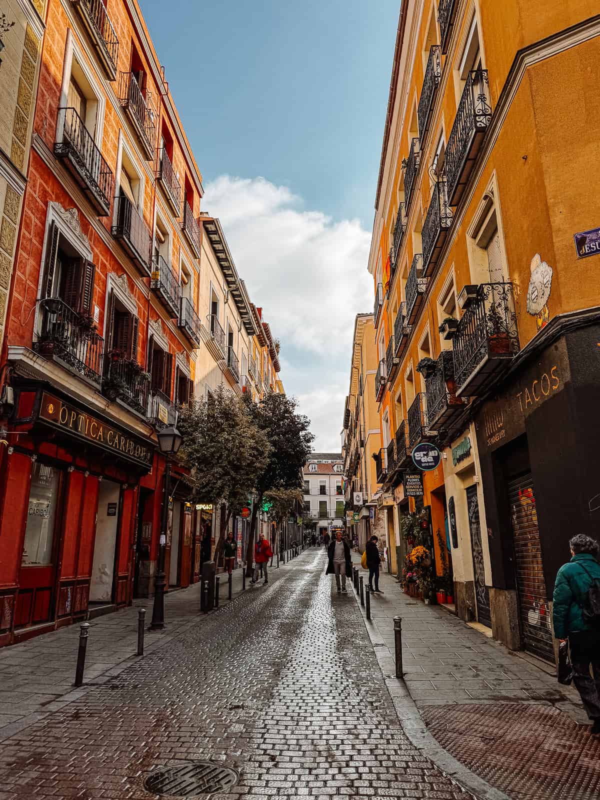 A picturesque view of a historic street in Madrid, with vibrant orange and yellow facades, ornate balconies, and pedestrians enjoying a sunny day.