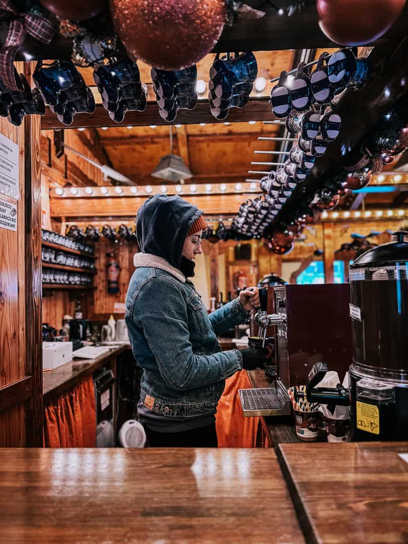 Market vendor in a hooded jacket pours a drink at a Vienna Christmas market, with rows of decorative glasses overhead.