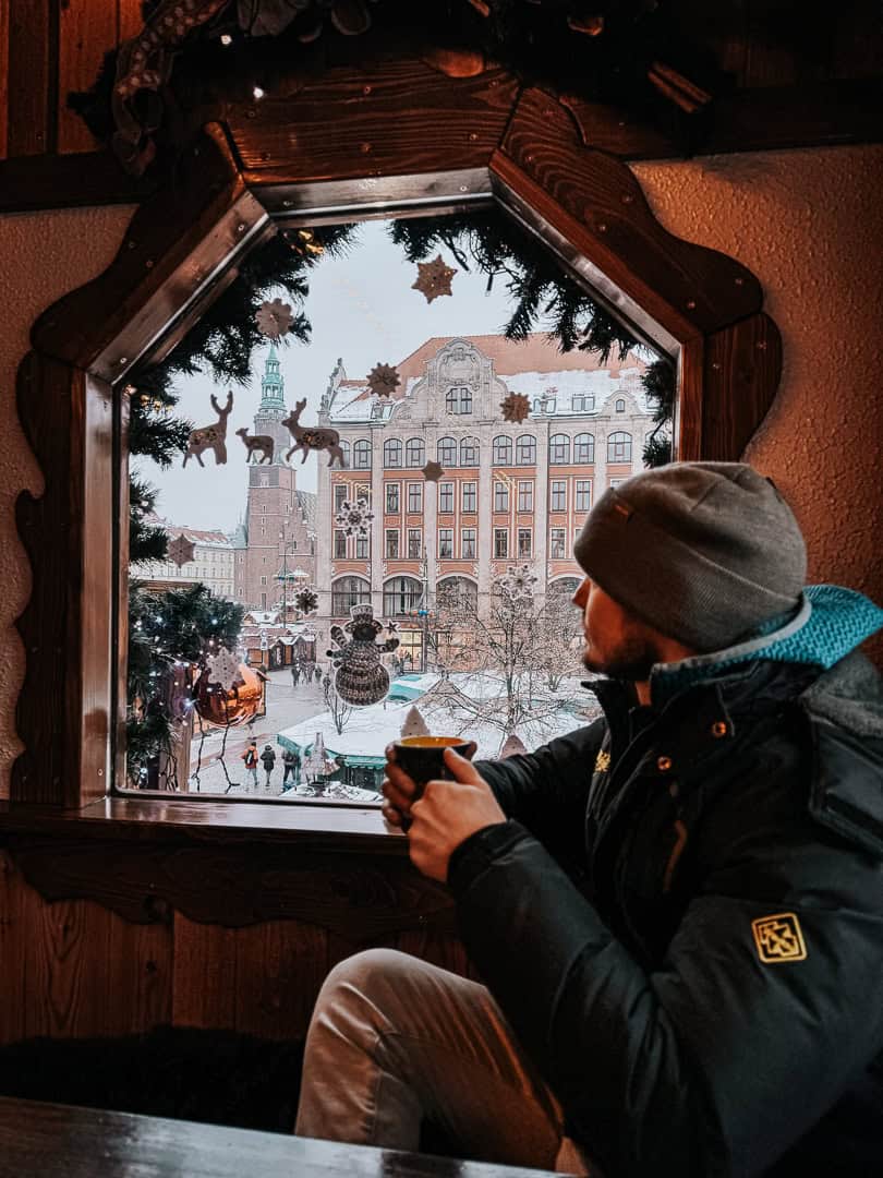 Man in a beanie enjoying a hot drink, looking out from a Christmas market stall with a picturesque view of Wroclaw's architecture