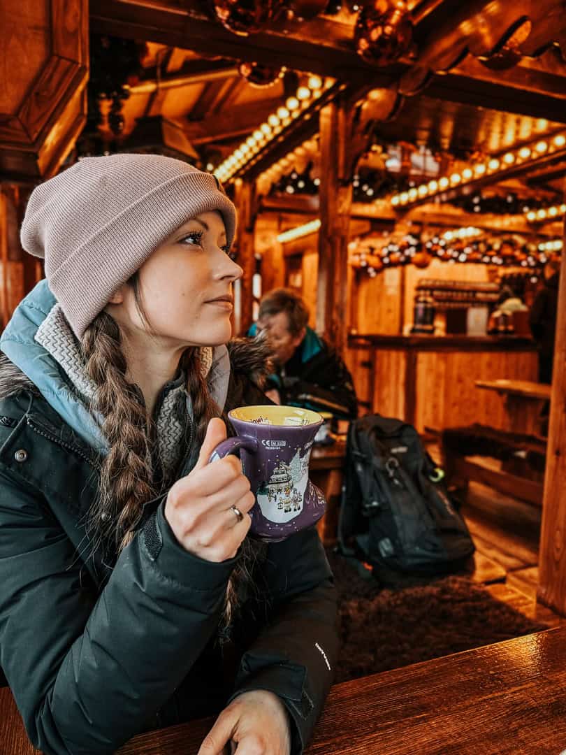 Woman in a beanie sipping a drink in a purple cup, with Christmas lights and market activity behind her