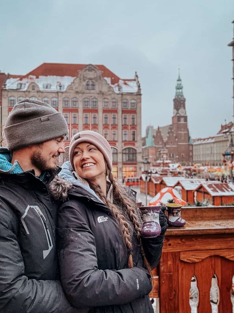 Couple enjoying mugs of mulled wine, smiling and embracing at a Wroclaw Christmas market, with historic buildings in the backdrop