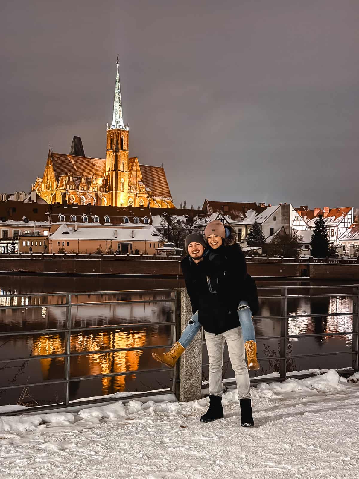 A smiling couple stands in the snow at night, with the illuminated Gothic architecture of Wroclaw’s Cathedral reflecting in the Oder River behind them