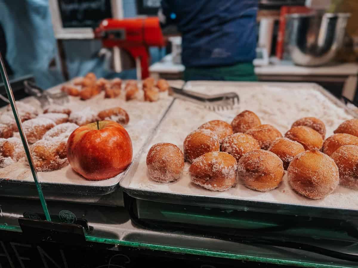 A fresh red apple sits prominently among trays of sugar-dusted doughnuts in a food stall, offering a colorful contrast to the golden brown treats.