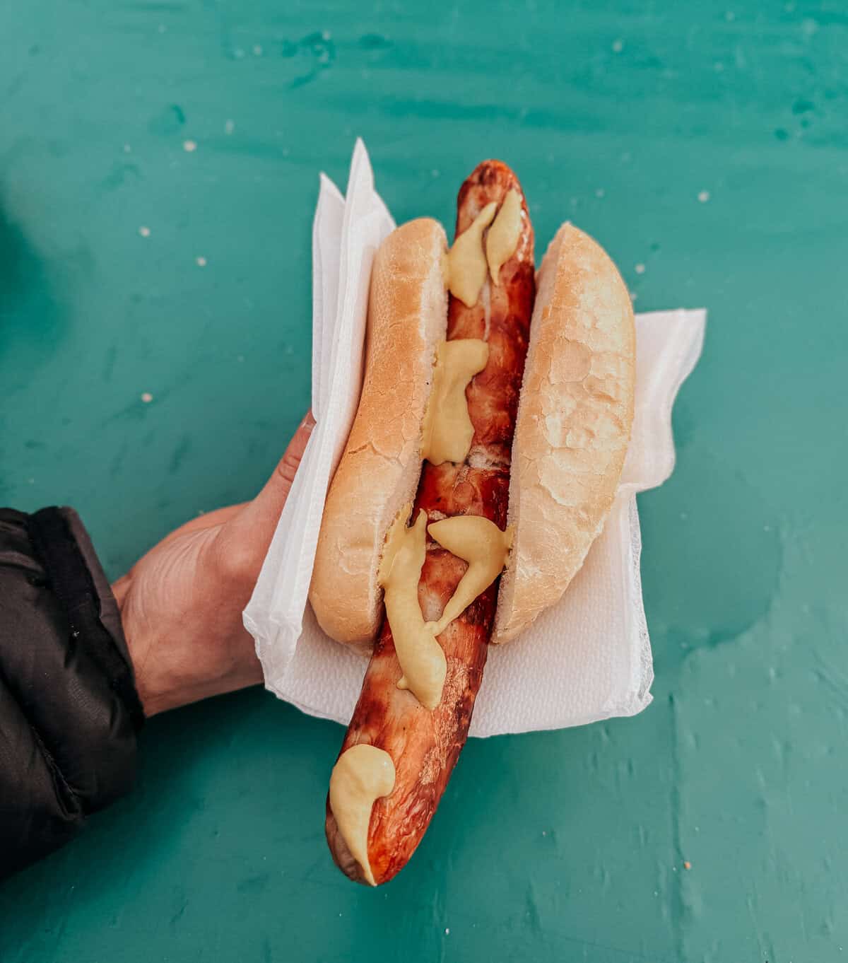 A hand holds a bratwurst sandwich with a crispy sausage topped with creamy mustard on a split white roll, set against a teal background