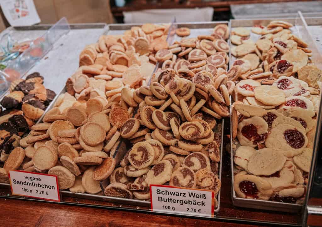 Trays filled with various traditional German cookies, including vegan options, with labels displaying prices, in a bustling market setting.
