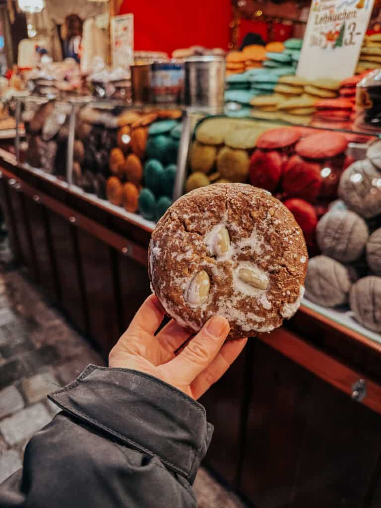 A hand holding a large Lebkuchen cookie decorated with almonds on top, with a market stall background filled with various goods.