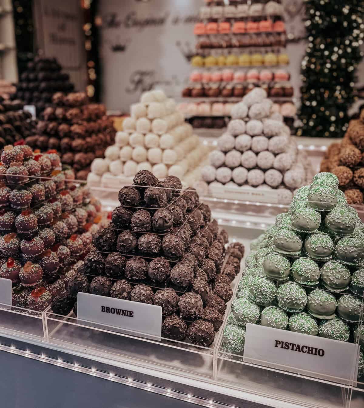 An enticing display of marzipan sweets arranged in pyramid shapes with various flavors like Brownie and Pistachio, each labeled and showcased against a backdrop of colorful macarons and a Christmas tree.