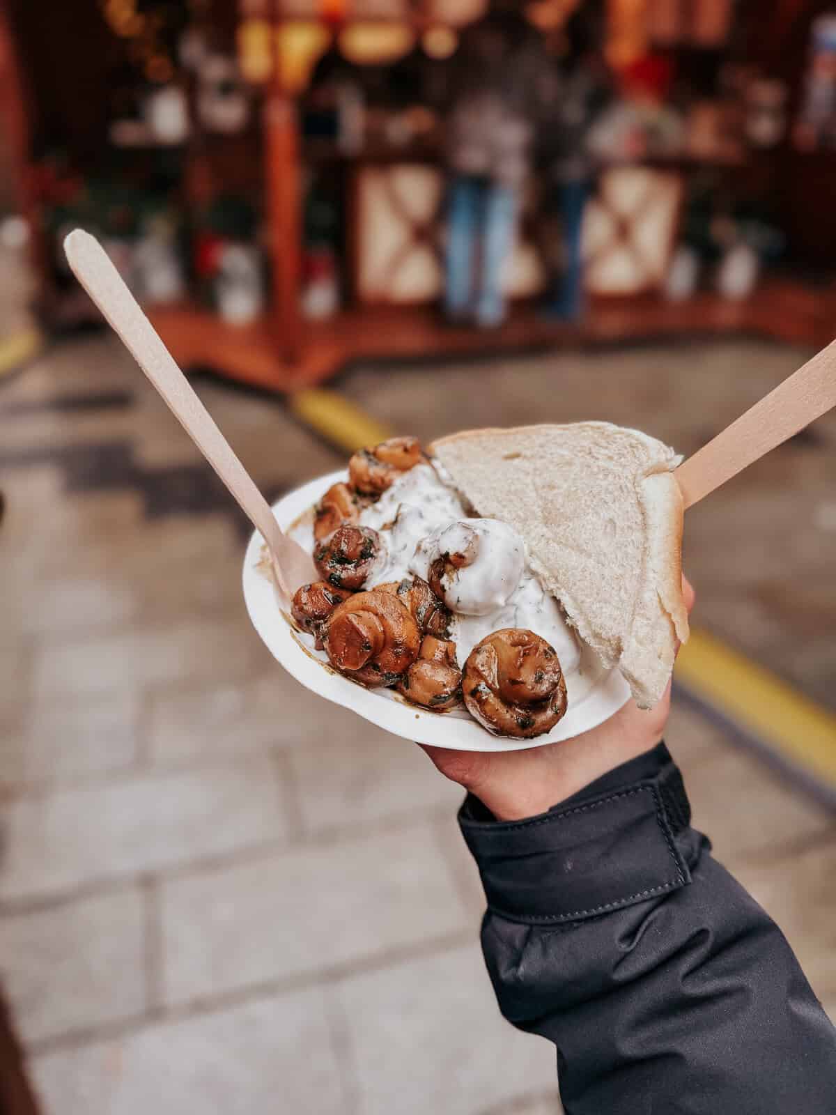 A hand holding a plate with roasted mushrooms topped with a creamy sauce served on a slice of bread, with street market stalls in the background