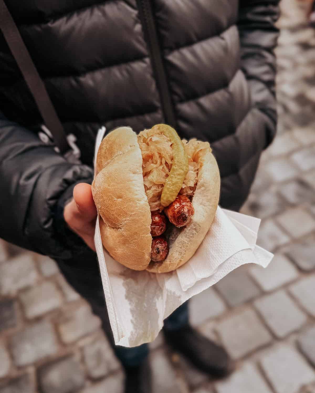 A person holding a Nuremberg sausage sandwich filled with small grilled sausages and sauerkraut, topped with mustard, in a crusty white bun.