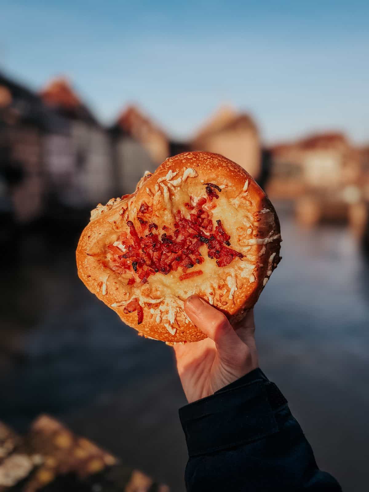 A person holding up a pretzel topped with melted cheese and red pepper strips against an old town backdrop, showcasing a creative street food snack