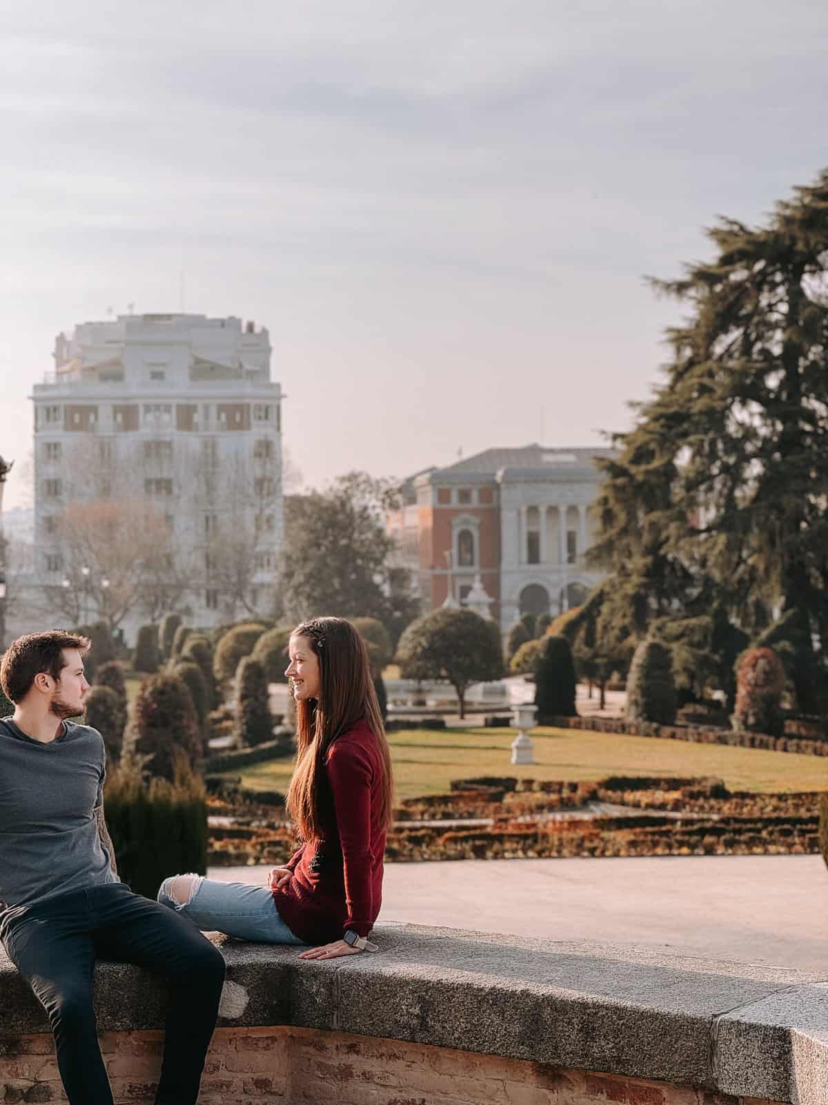 A man and a woman sit facing each other on a stone wall, engaging in a casual conversation with a serene, landscaped garden and classic European architecture in the background, all bathed in the soft glow of the afternoon sun