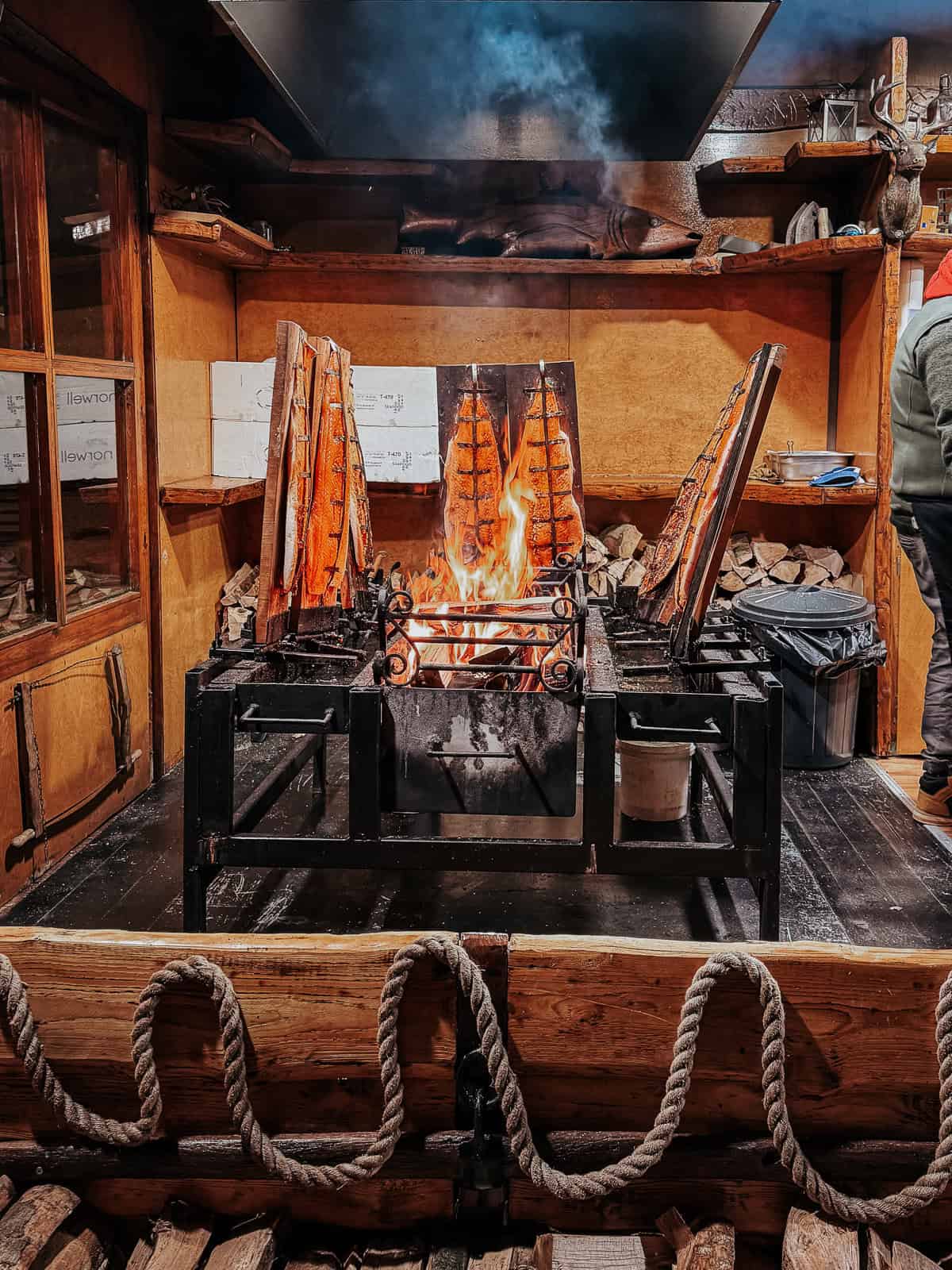 Fresh salmon fillets cooking on a unique vertical open fire grill, surrounded by a rustic wooden frame and smoke extraction setup at a market stall.