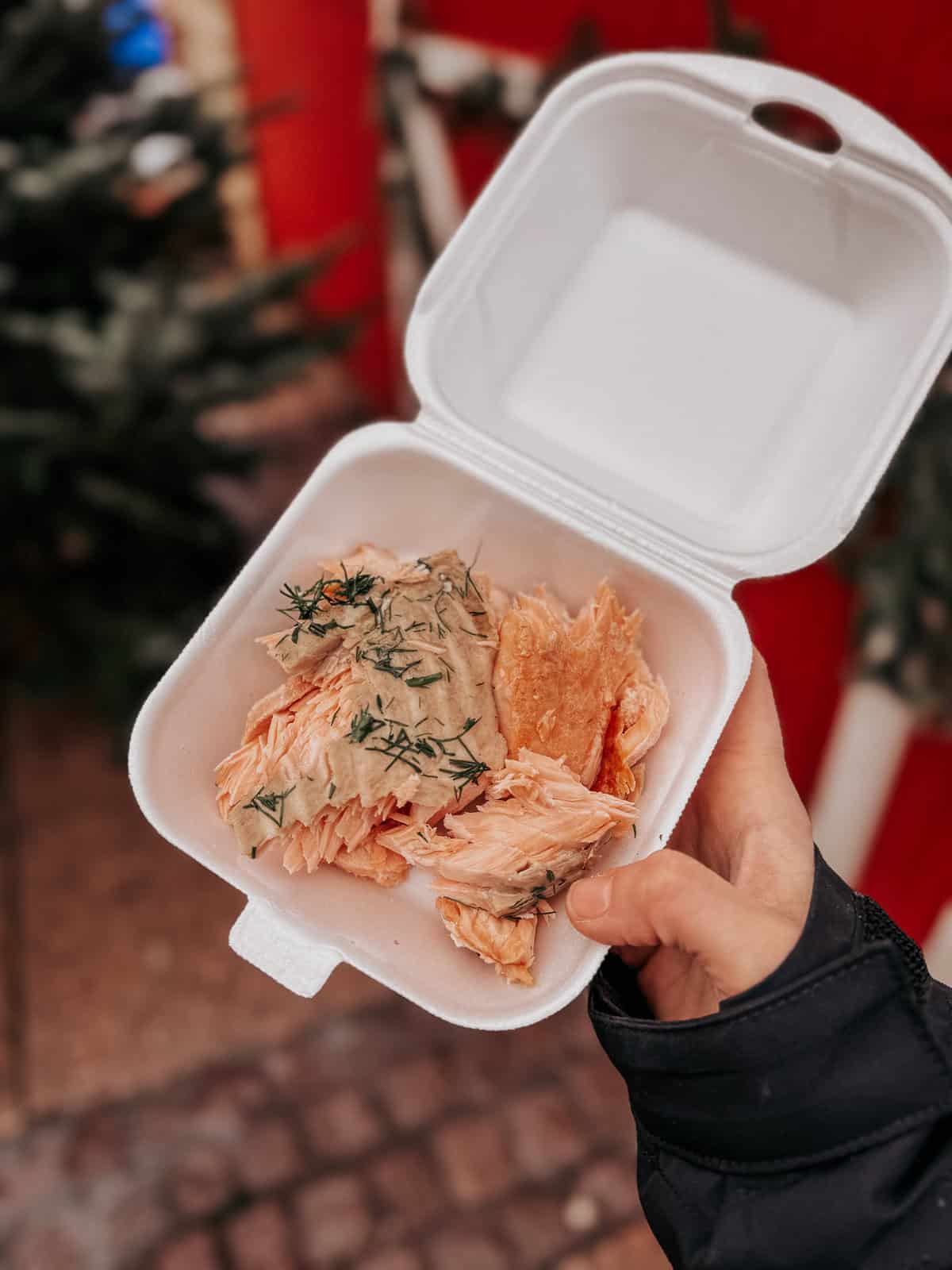 Close-up of a takeout container held in hand, filled with flaked salmon seasoned with fresh herbs, emphasizing a quick and savory street food option.