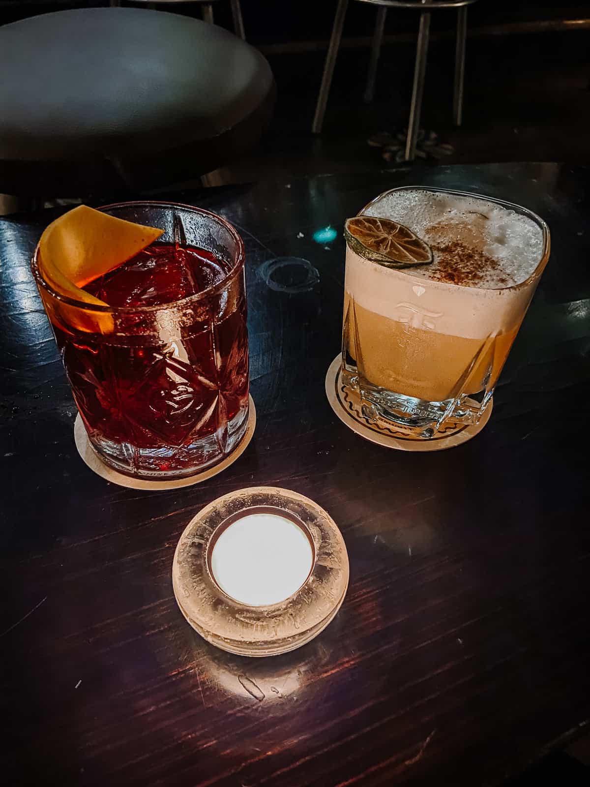 Two artisanal cocktails sit invitingly on a dark wood table; one is a deep red with a citrus peel garnish, and the other is a creamy, frothy concoction topped with a dried citrus slice, both casting a cozy glow next to a round, lit candle.