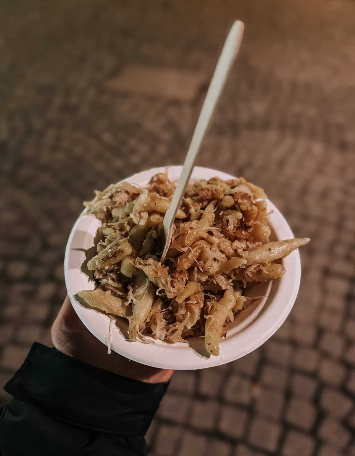A hand holding a plate filled with Schupfnudeln mixed with sauerkraut and sprinkled with crispy bacon bits, served at a night market.