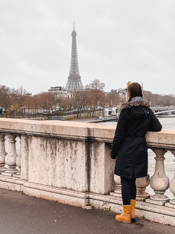  A woman in winter attire contemplates the Eiffel Tower from a stone bridge over the Seine River in Paris, embodying the allure of solo travel in Europe with the iconic iron lattice tower in the backdrop.