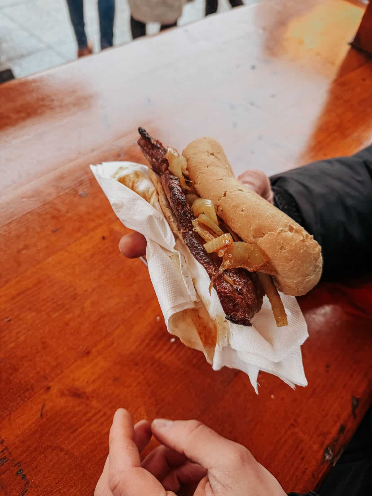 A street food vendor handing over a steak sandwich loaded with grilled steak, caramelized onions, and pickles in a crusty baguette, wrapped in a napkin