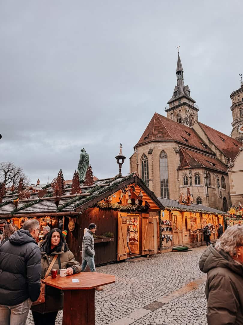 A daytime scene of the Stuttgart Christmas market showing visitors near wooden stalls, with the Old Castle's tower and a statue in the background.