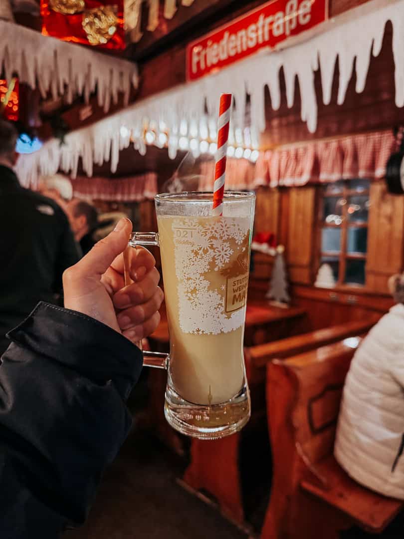 A person holding a glass of hot beverage with a striped straw, with a cozy Christmas market stall blurred in the background.