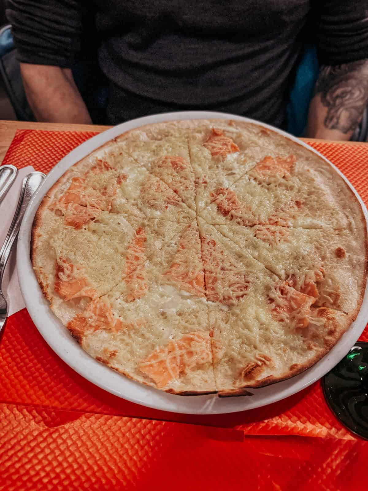 A large, freshly baked Tarte Flambée with a thin crust topped with cream, onions, and salmon, served on a white plate