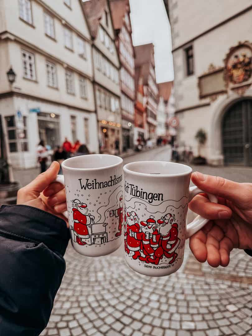 Two hands holding special edition Christmas mugs with Santa Claus motifs, against the backdrop of Tübingen's picturesque half-timbered houses.