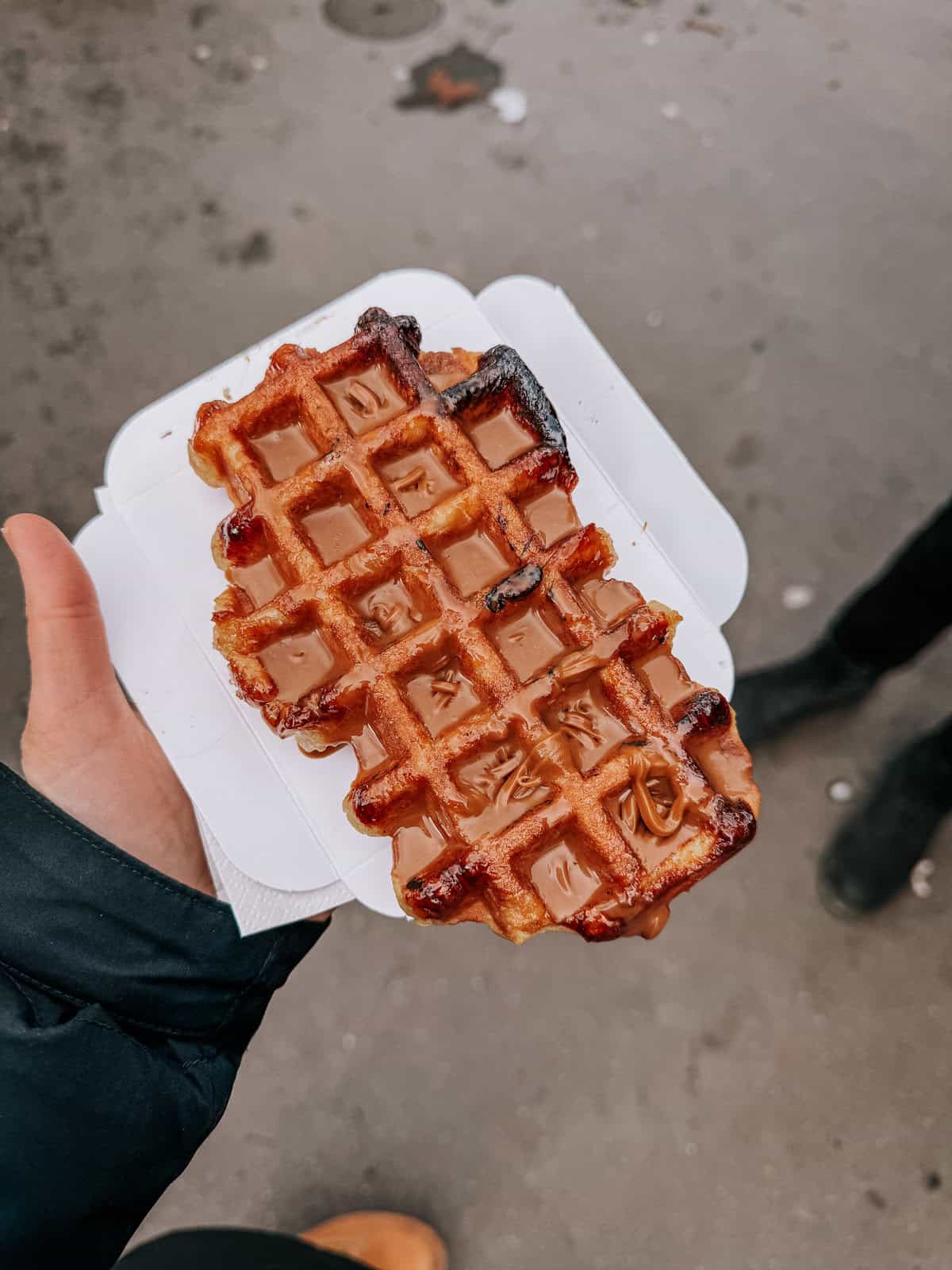 A close-up of a caramel-topped waffle, held in a hand with the city street blurred in the background, showcasing a gooey caramel drizzle in the grid of the waffle