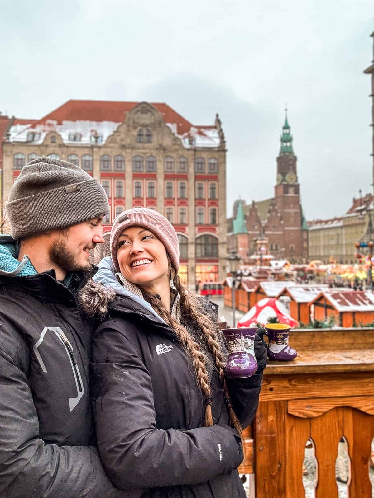 A smiling couple, holding mulled wine in festive cups, enjoys the Christmas market in Wroclaw, with the colorful facades of the town's historic buildings and a dusting of snow in the background