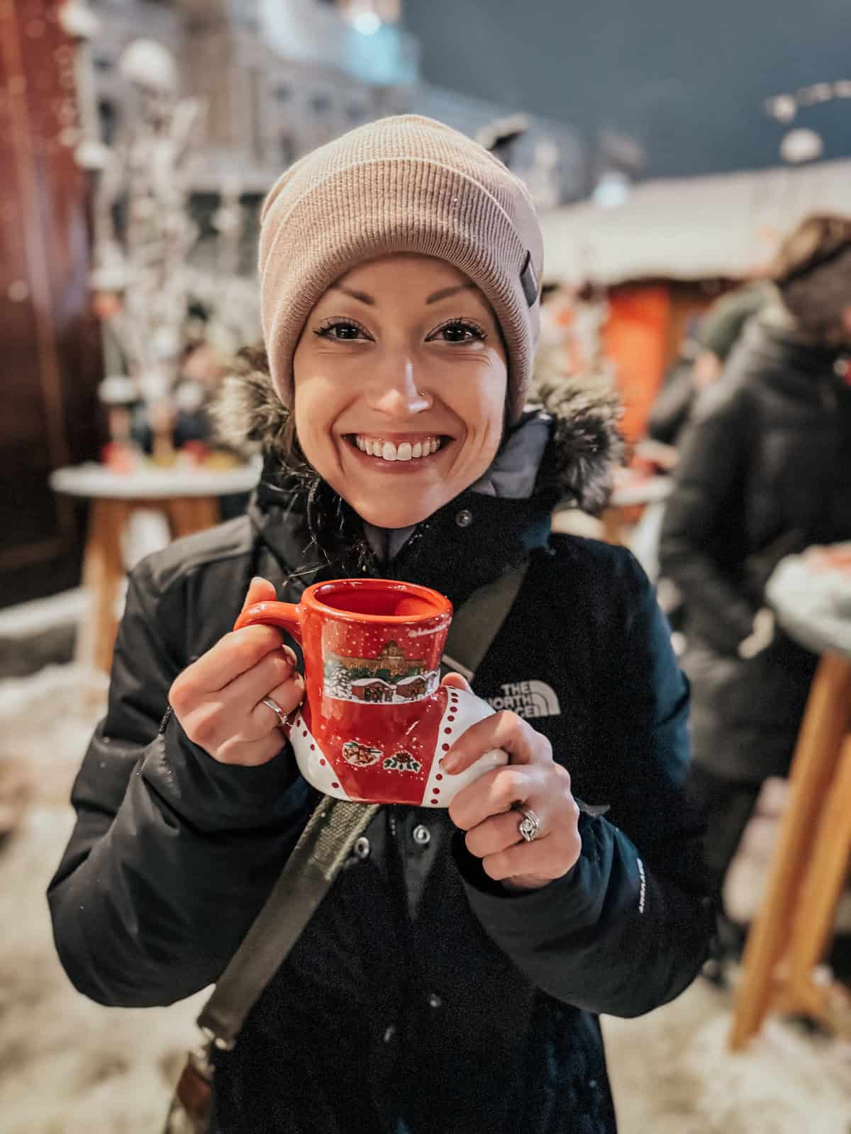 a smiling woman in a pink hat holding a red boot mug at a christmas market