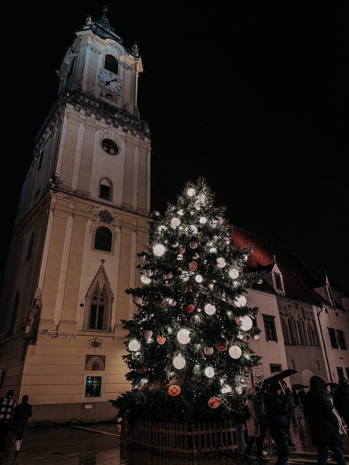 A majestic Christmas tree illuminated with white lights, standing before a tall, ornate church tower in a lively night setting, with people milling around the square.