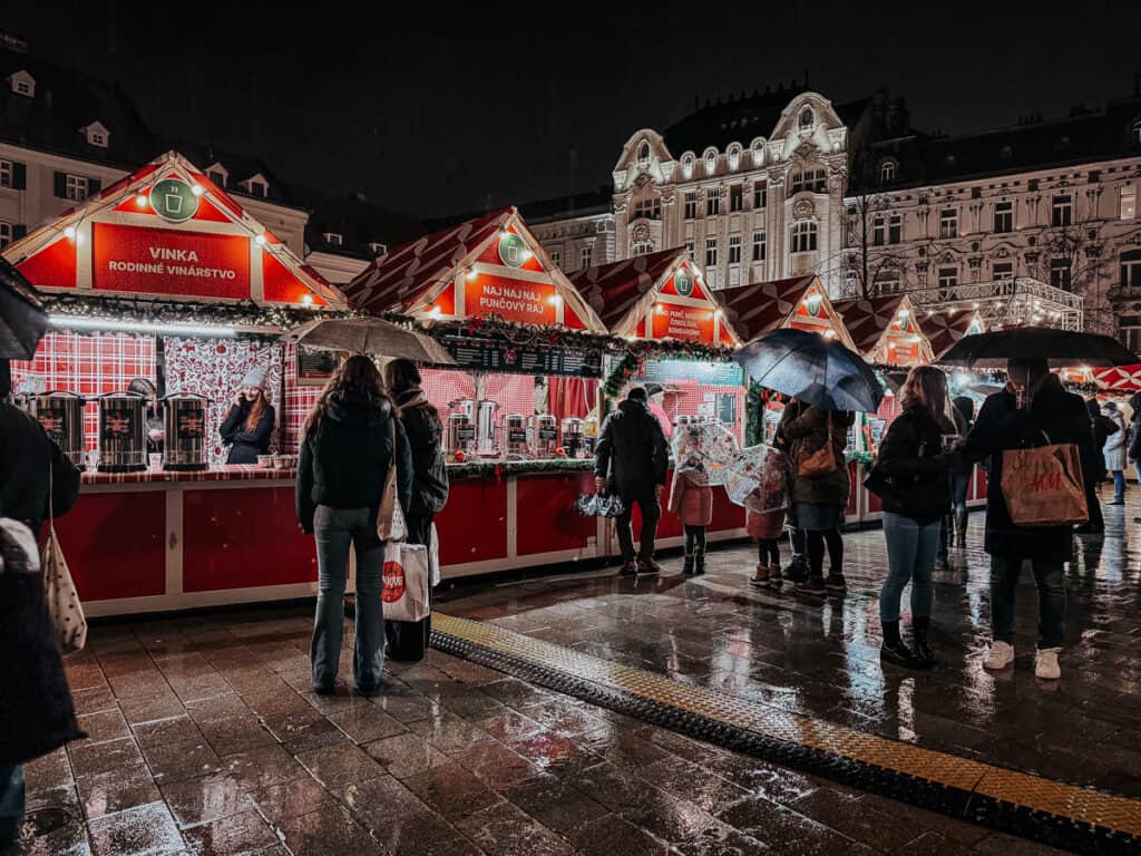 A bustling night market in Bratislava with festive lighting and rain-soaked pavements, people shopping under umbrellas.