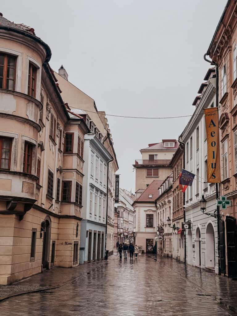 A historical, cobblestone street in Bratislava, lined with old European buildings under a cloudy sky, reflecting a wet surface.
