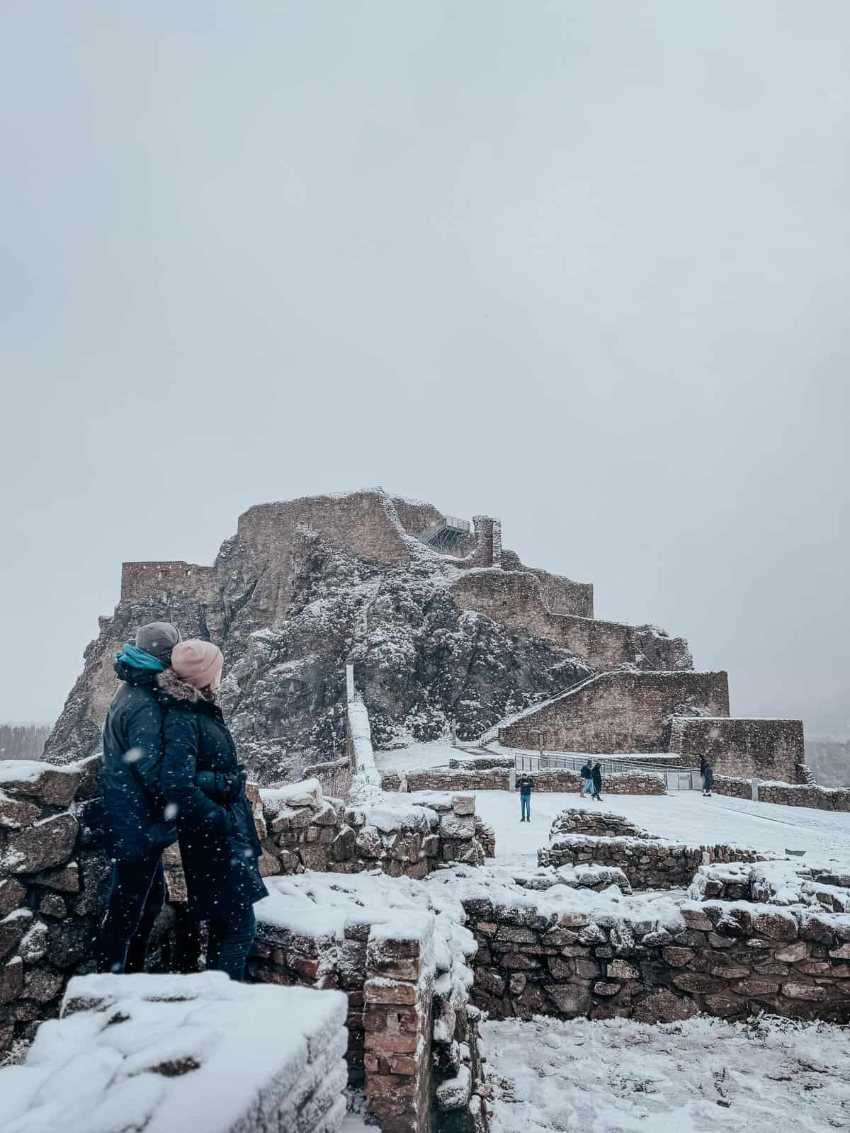 Two people in winter attire gaze at a snowy hilltop castle ruins, with snowflakes falling around them and other visitors in the distance.