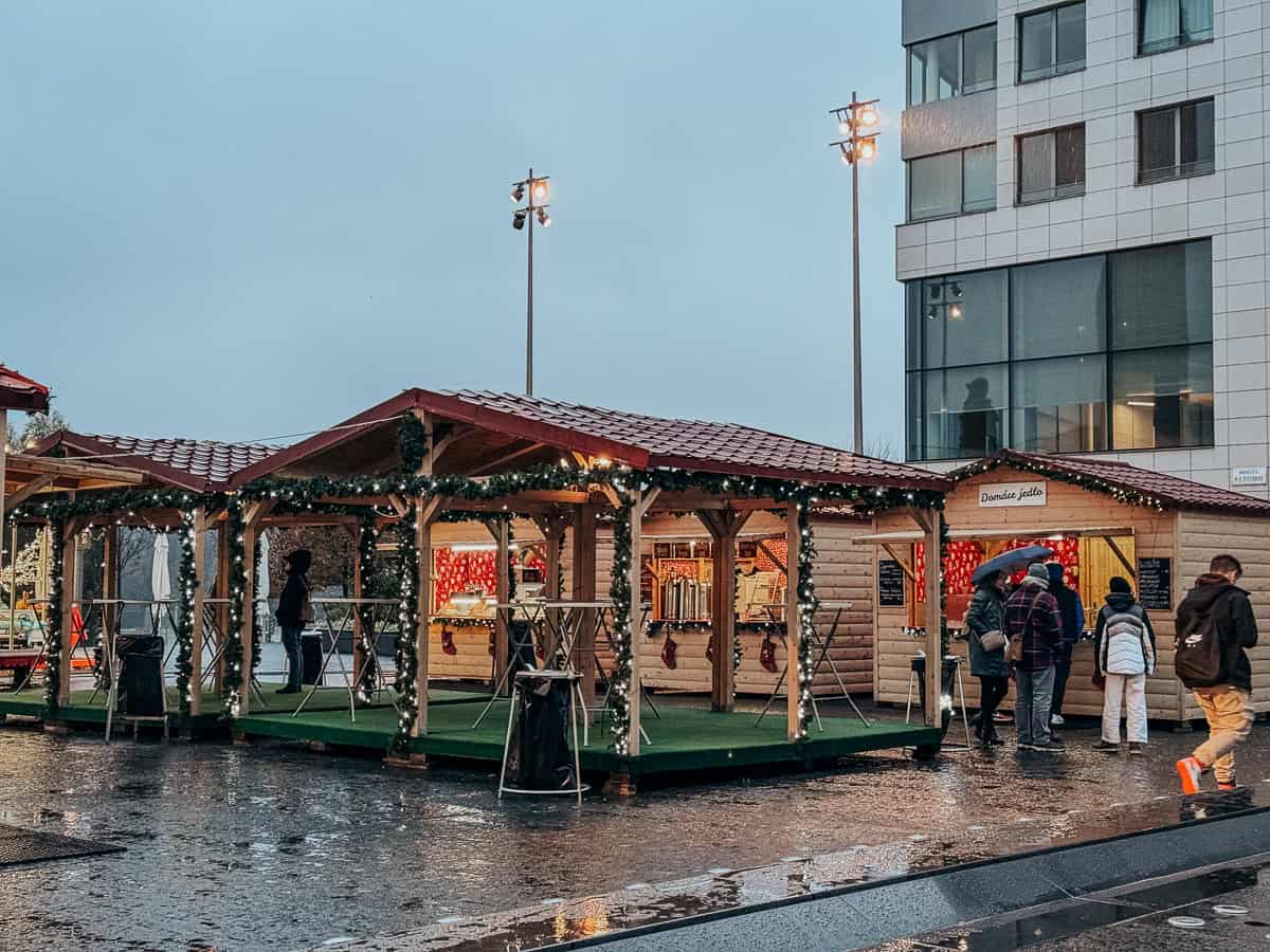 An outdoor Christmas market scene with several decorated wooden stalls under shelter, reflecting a wet pavement and festive lights, bustling with holiday shoppers.