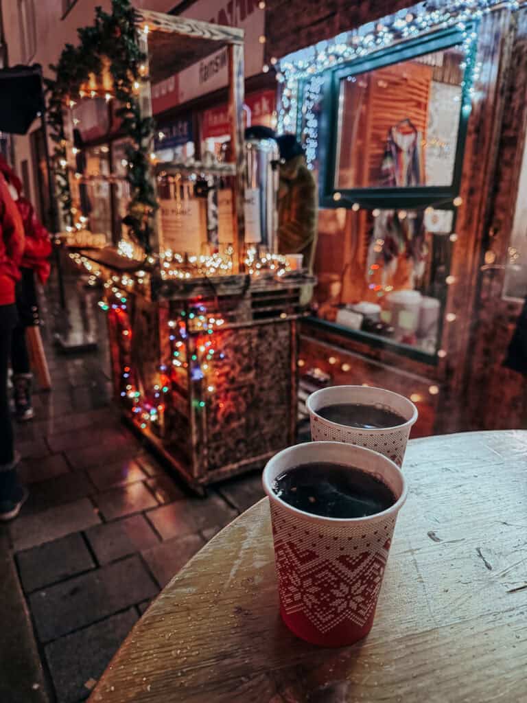 Two cups of mulled wine on a wet tabletop with festive decorations and a market stall in the background, showcasing a cozy, holiday market experience.