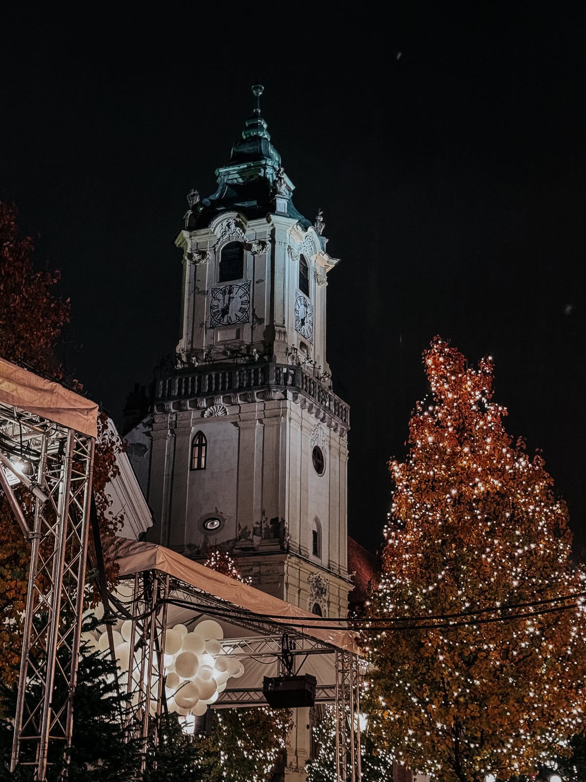 A close-up of a church tower lit against the night sky, framed by festive decorations and glowing trees, capturing the serene ambiance of a Christmas market.