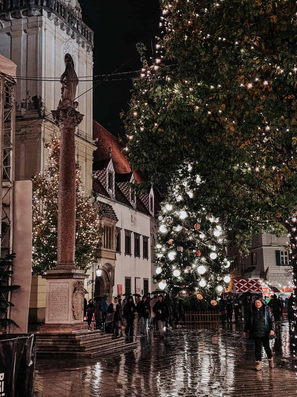 A picturesque night scene in a square with a historical statue, a large Christmas tree, and a crowd of visitors under twinkling lights and a wet pavement reflecting the festive atmosphere.