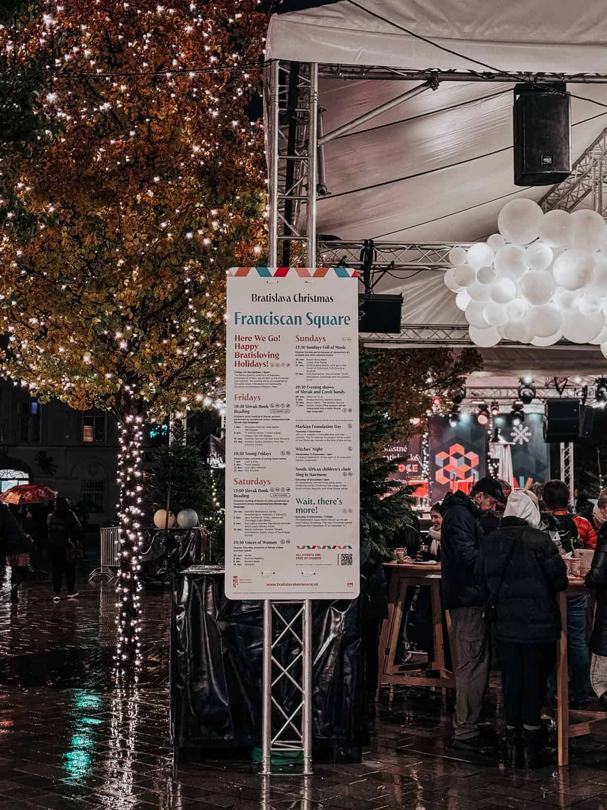 A detailed view of a Christmas market event sign at Franciscan Square in Bratislava, surrounded by lit-up trees and people enjoying the market under sheltered areas in rainy weather.