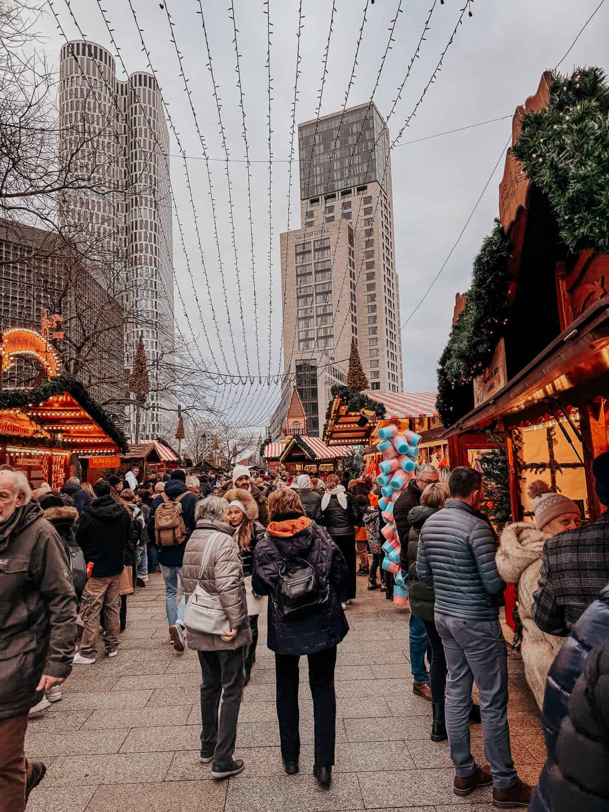 A Christmas market set against a backdrop of modern high-rise buildings, adorned with festive decorations and strings of lights overhead. Crowds of shoppers explore various stalls offering holiday treats and goods