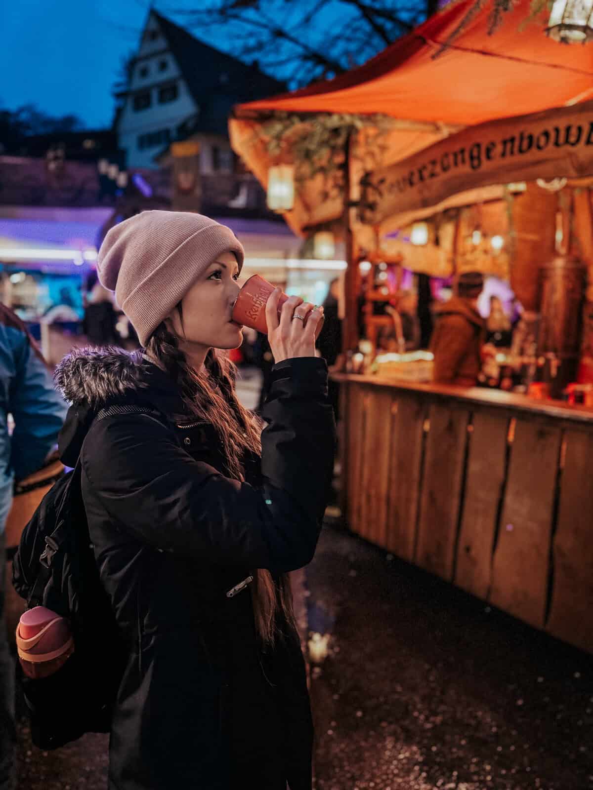 a woman enjoying a cup of mulled wine, with market stalls and festive lights in the background.