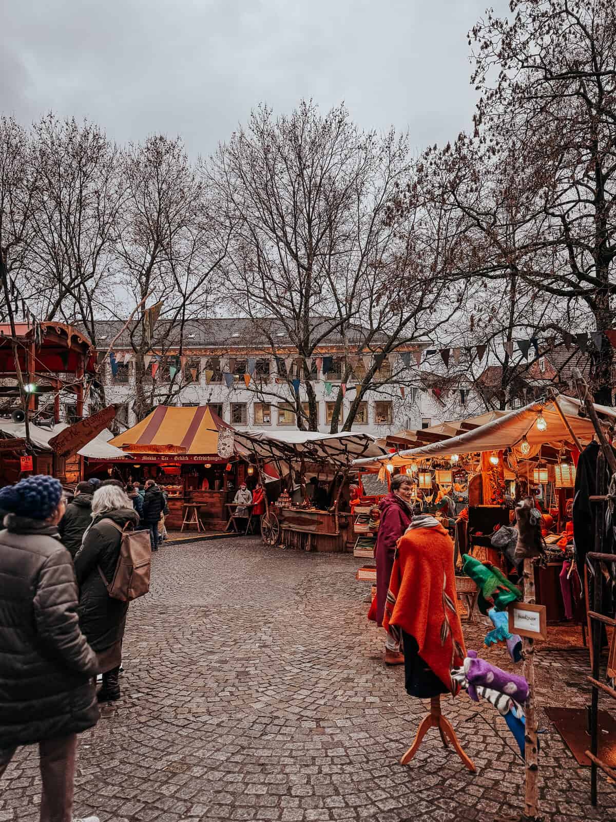 a busy market scene with various stalls, medieval banners, and a crowd of visitors enjoying the atmosphere.