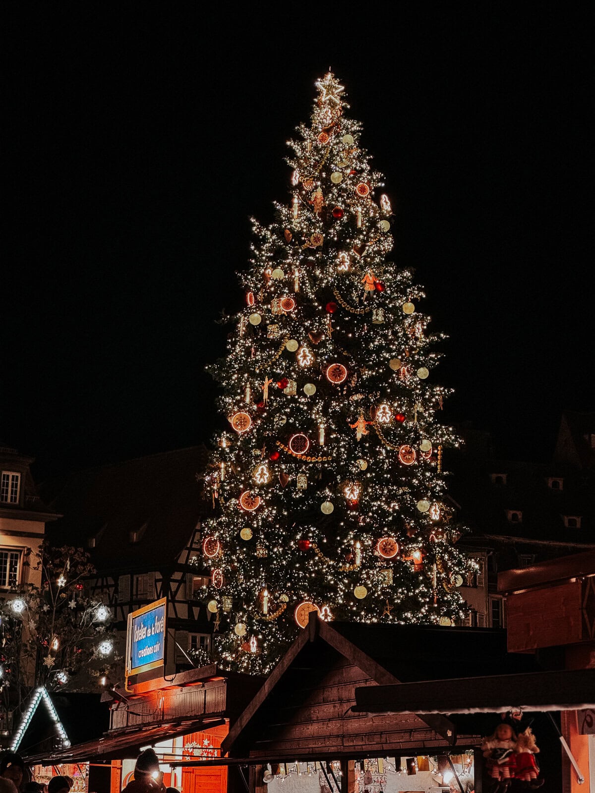 A large Christmas tree beautifully adorned with lights and ornaments, standing in a market square surrounded by half-timbered houses and festive stalls.