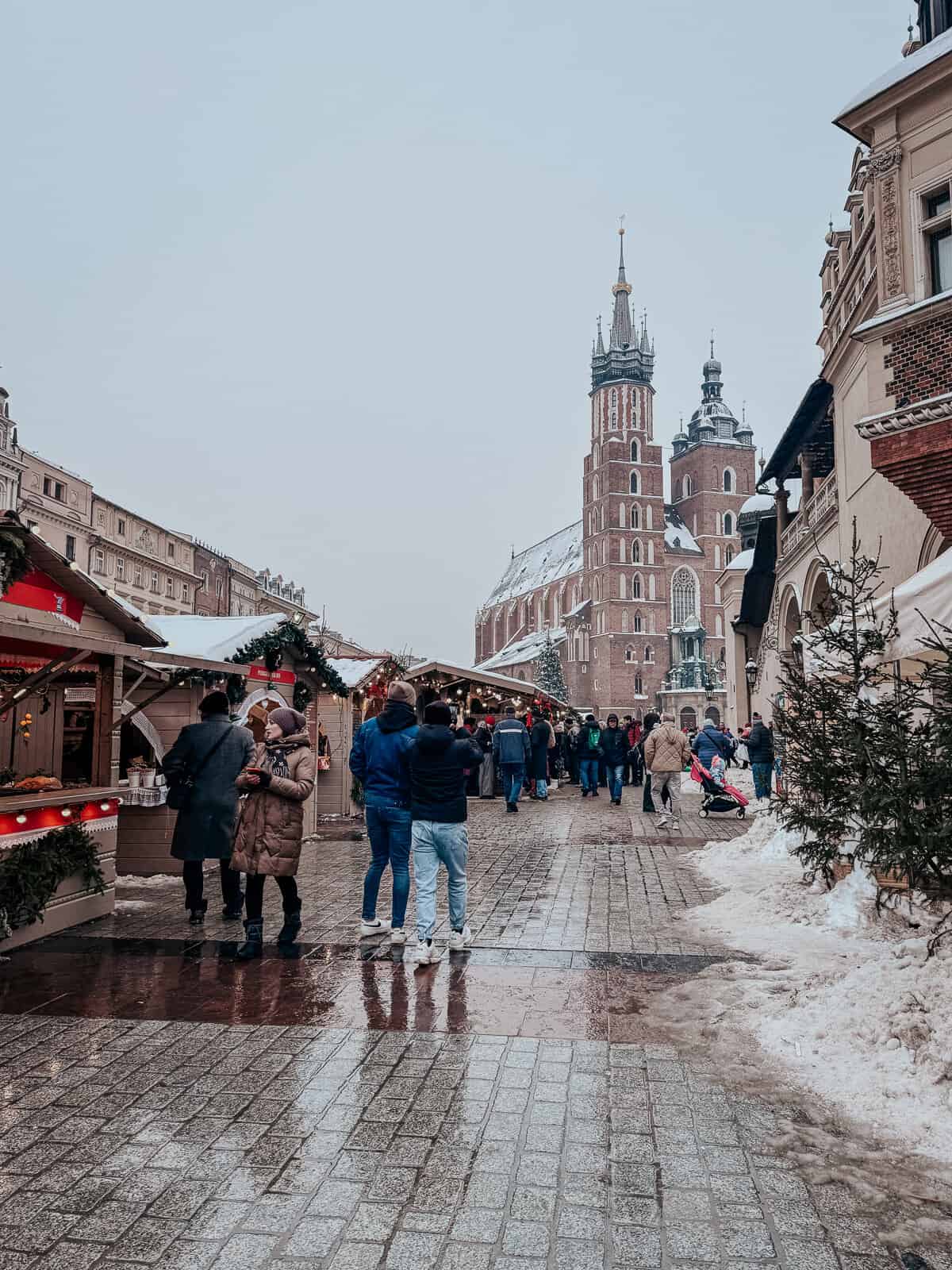 People walking through a snowy Christmas market street, with red-roofed stalls on either side selling holiday items, with the iconic red brick Gothic St. Mary's Basilica in the distance under a gray sky.