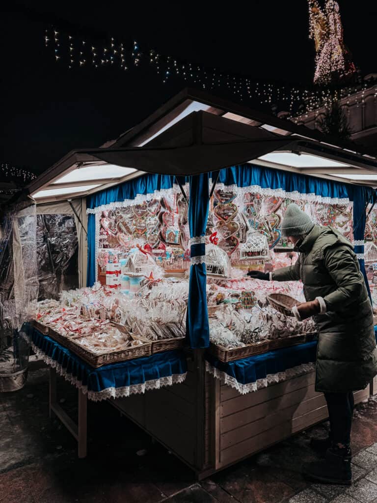 A market stall at a Christmas fair selling intricately decorated gingerbread cookies, covered by a blue canopy, with a bundled-up vendor arranging the displays, set against a dark evening sky lit by festive lights.