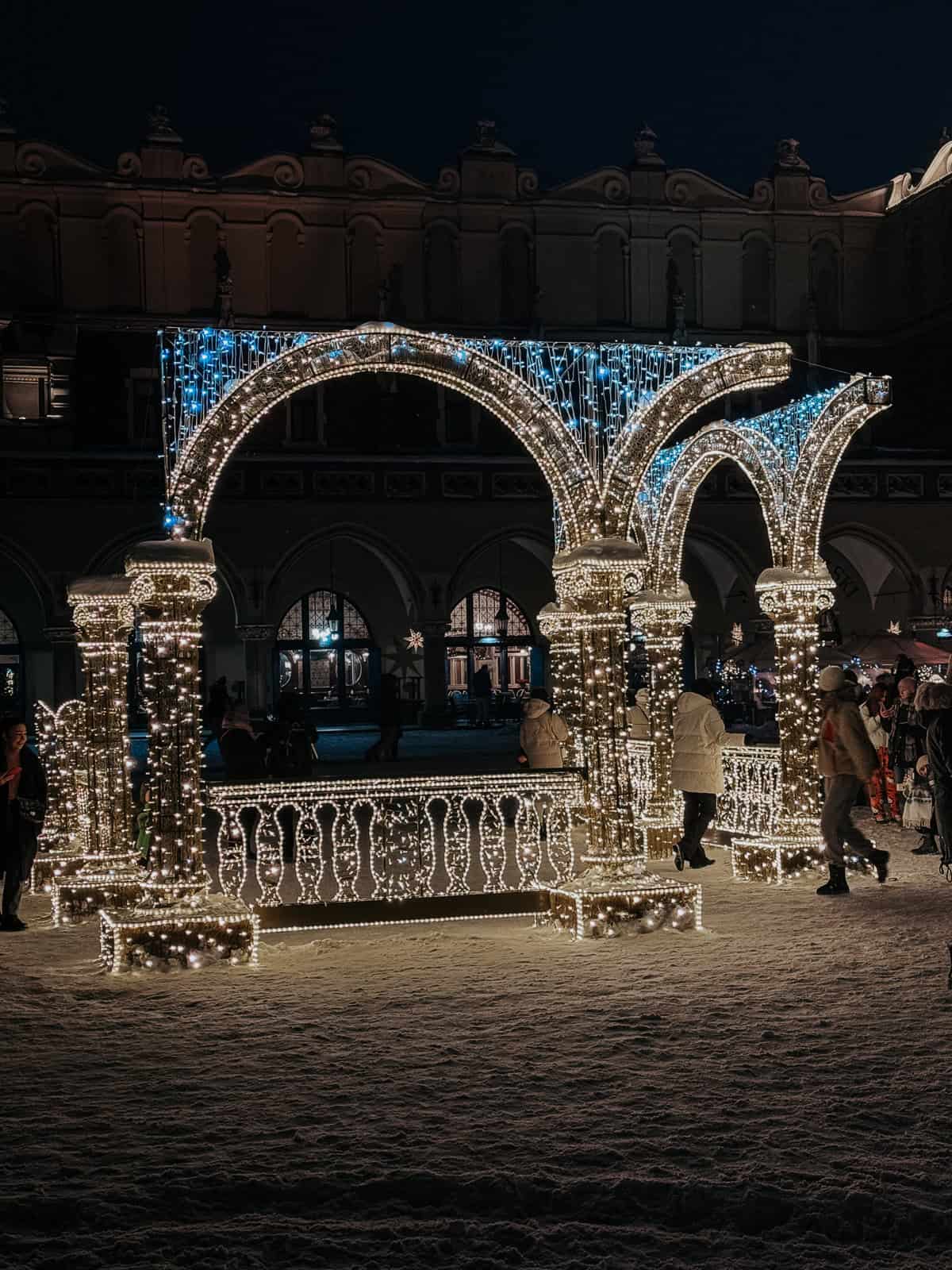 A festive, illuminated archway adorned with blue and white lights creating a gateway, surrounded by snow-covered ground at a night-time Christmas market, with people milling about and historic buildings in the background.