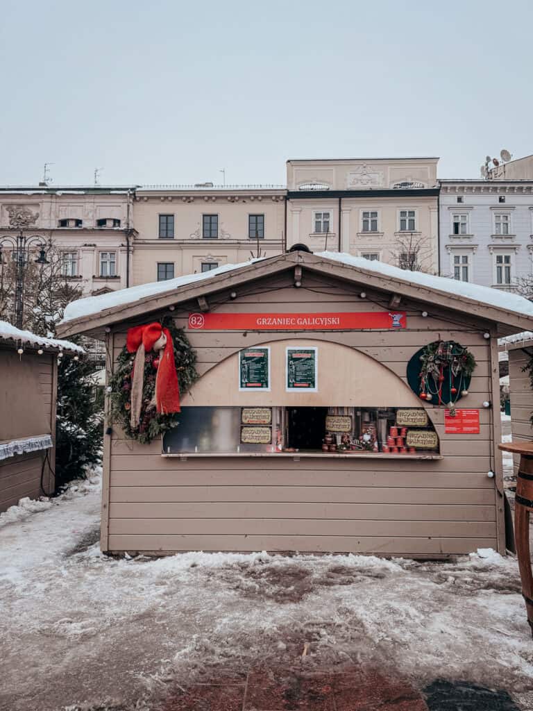 A wooden food stall decorated with red accents and holiday greens under a dusting of snow, displaying signs for 'Grzaniec Galicyjski' which is a traditional Polish spiced wine, set against a backdrop of historic pale-colored buildings
