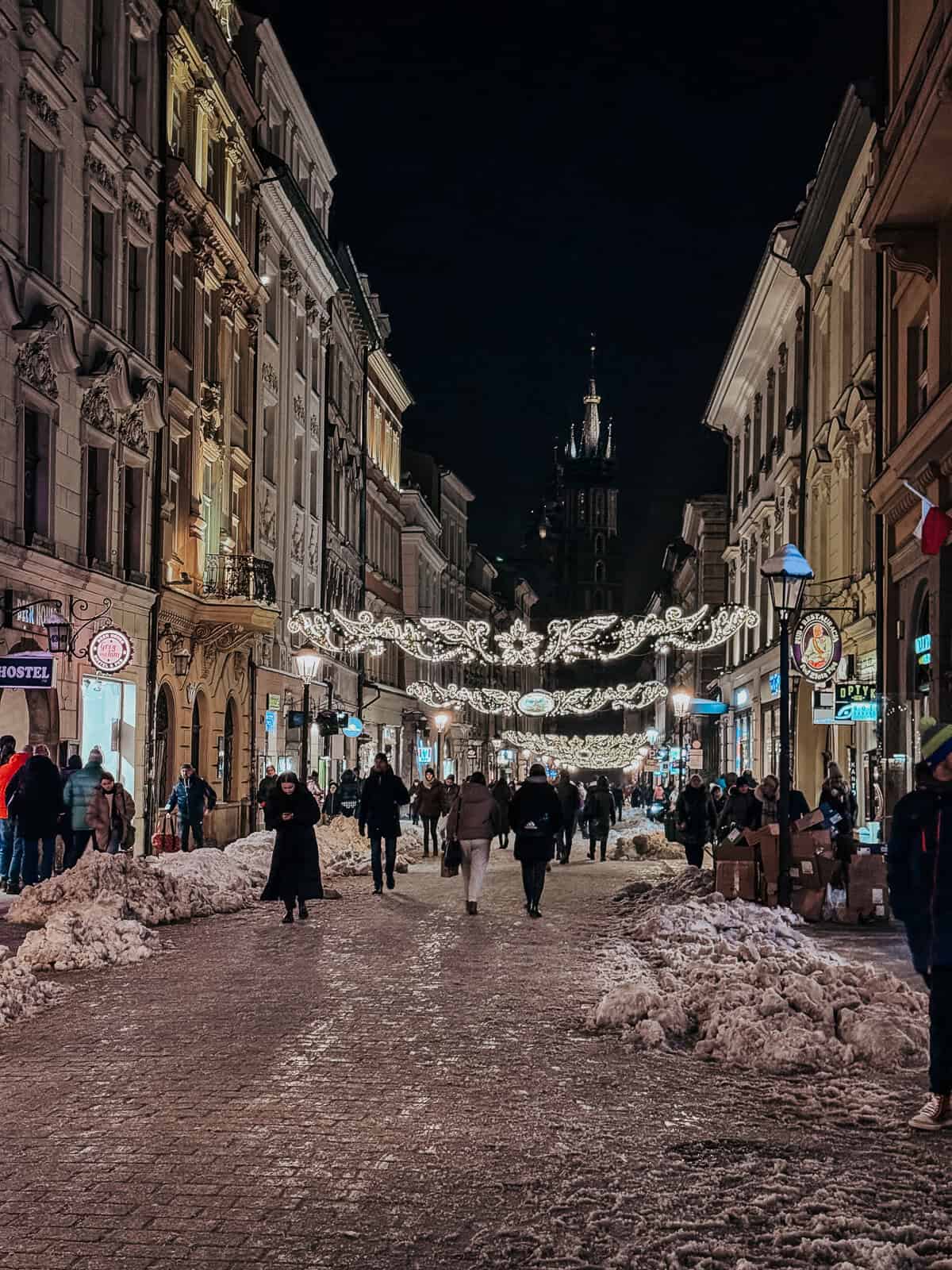 A bustling street view at night in Krakow during the Christmas season, decorated with festive lights and filled with people walking, with snow piled up along the sides